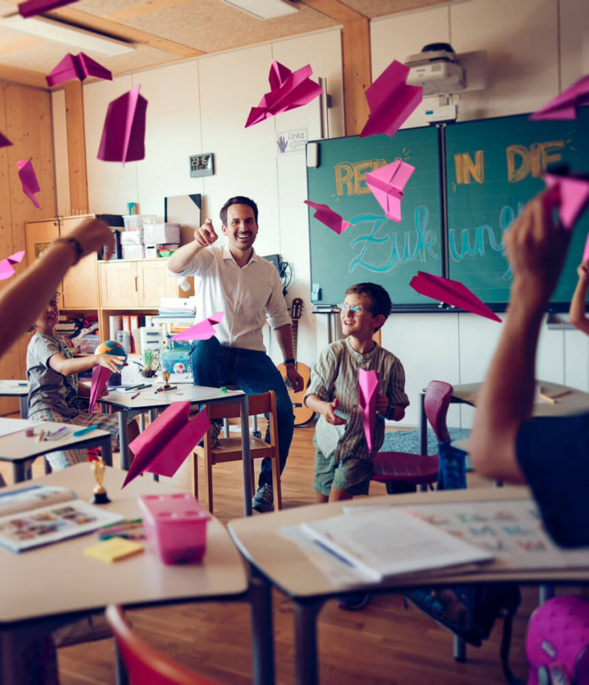 Foto von Bundesminister Christoph Wiederkehr mit Kindern in einem Klassenzimmer, es werden Papierflieger geworfen