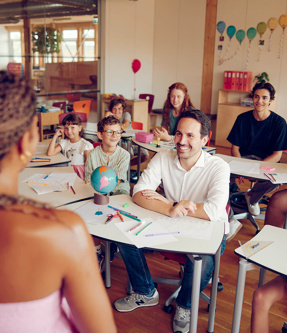 Bundesminister Christoph Wiederkehr sitzt zwischen Kindern im Klassenzimmer, Blick Richtung Tafel