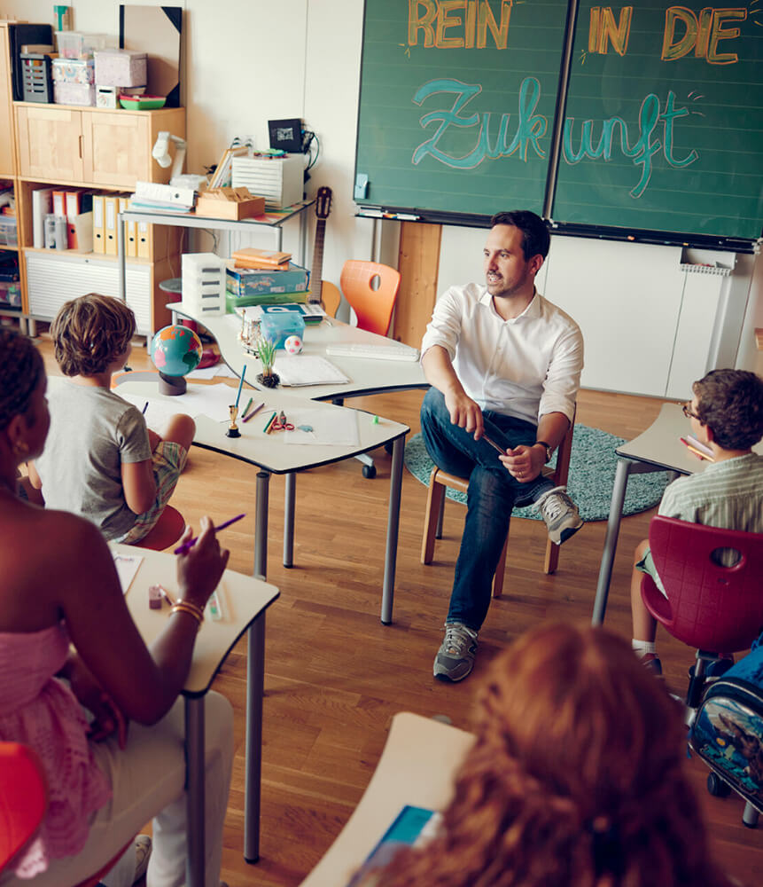 Bundesminister Christoph Wiederkehr sitzt im Klassenzimmer mit Rücken zur Tafel und spricht mit Kindern