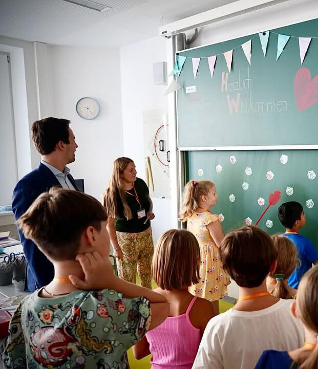 Bundesminister Christoph Wiederkehr steht mit Kindern im Klassenzimmer und schaut auf die Tafel wo Herzlich Willkommen geschrieben steht