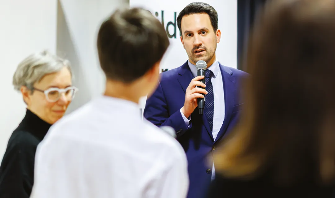 Christoph Wiederkehr im Anzug spricht mit Mikrofon vor einer kleinen Gruppe von Zuhörenden in einem Innenraum.
