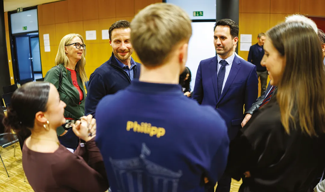 Christoph Wiederkehr im Gespräch mit einer Gruppe von Schüler:innen und Erwachsenen in einem Innenraum, lockere Interaktion in kleiner Runde.